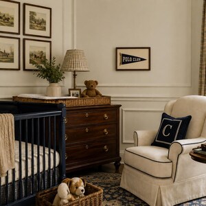 Op de afbeelding: Een kinderkamer met een donkerblauw ledikant met een beige kabelgebreide deken, een donkerhouten ladekast met een geruite lamp en teddybeer, een cr&egrave;mekleurige fauteuil met een donkerblauw kussen met de letter C en een Polo Club wimpel aan de muur.