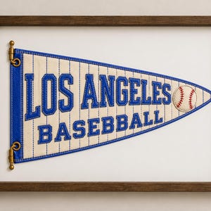 以下が含まれることがあります： クリーム色の生地に青いピンストライプが施され、青い刺繍で「LOS ANGELES BASEBALL」と書かれた額装済みの三角形のペナント。ペナントの右側には、赤いステッチが入った白い野球ボールの小さなグラフィックが描かれています。