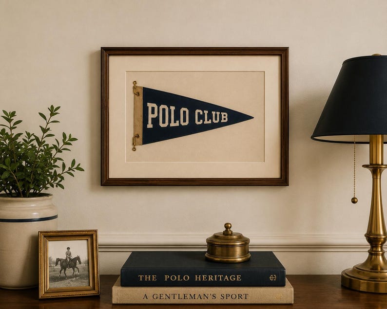 Op de afbeelding: Een ingelijste marineblauwe driehoekige wimpel met de tekst POLO CLUB in witte letters, gemonteerd op een cr&egrave;mekleurige passe-partout in een donkerhouten lijst. De wimpel is aan de linkerzijde bevestigd met messing beslag en toont een sportief ontwerp.