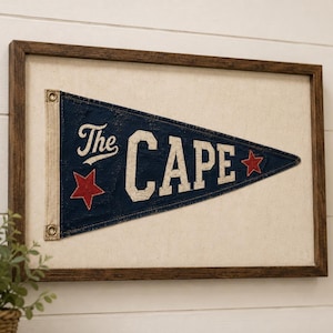 May include: A framed wall art piece featuring a triangular navy blue pennant with the text The CAPE in white lettering. The pennant includes two red stars and is mounted on a light-colored fabric background within a dark brown wooden frame.