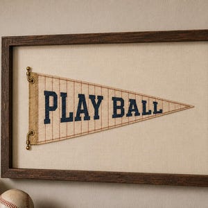 May include: A framed wall art piece featuring a triangular beige pennant with thin red vertical pinstripes and the text PLAY BALL in dark blue block letters. The pennant is attached to a dark brown wooden frame with small brass hardware.