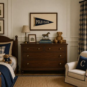 Op de afbeelding: Een slaapkamer met een donkerhouten ladekast, een bed met marineblauw beddengoed en een kussen met het label CHARLIE, een cr&egrave;mekleurige fauteuil met een marineblauw kussen met de letter C, een ingelijste Polo Club wimpel, paardenprints en een beer.