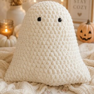 May include: A cream-colored, triangular-shaped plush toy crafted with a textured crochet stitch. The object features two small, round, black plastic eyes positioned near the top, giving it the appearance of a ghost figure.