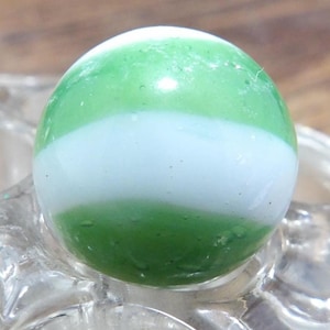 Peut inclure: Une bille en verre sphérique présentant une surface lisse avec des bandes horizontales alternées de verre blanc opaque et de verre vert translucide. La bille a une finition brillante et une forme arrondie claire qui reflète la lumière.