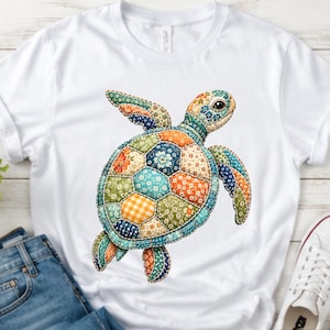 May include: A white short-sleeved t-shirt featuring a large graphic of a sea turtle. The turtle's shell and body are composed of various colorful patchwork fabric patterns, including floral, gingham, and geometric designs in shades of blue, orange, and green.