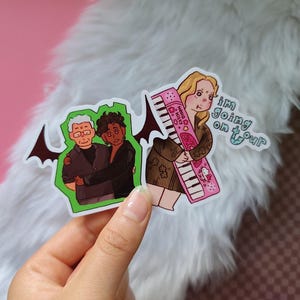 Könnte beinhalten: Zwei gestanzte Vinyl-Aufkleber. Ein Aufkleber zeigt zwei Personen, die sich vor einem hellgrünen Hintergrund mit schwarzen Fledermausflügeln umarmen. Der andere zeigt eine Person mit einem rosa Hello Kitty Keyboard und dem Text "im going on tour".