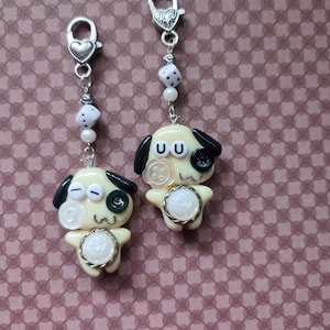 May include: Two silver-toned metal lobster clasp charms featuring pale yellow cartoon dog figures. Each dog has black ears, a button eye, a white floral button on the cheek, and a white circular belly accent. The charms include white dice beads.