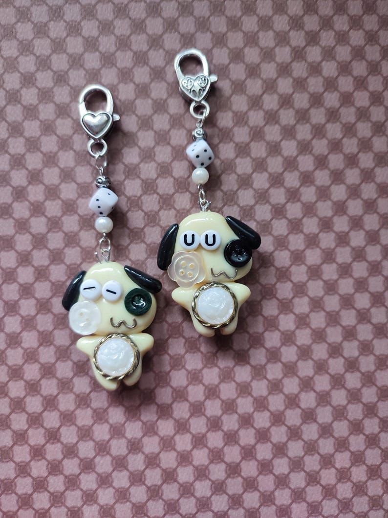 May include: Two silver-toned metal lobster clasp charms featuring pale yellow cartoon dog figures. Each dog has black ears, a button eye, a white floral button on the cheek, and a white circular belly accent. The charms include white dice beads.
