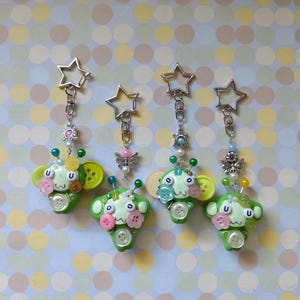 May include: Four green creature keychains featuring star-shaped metal clasps, silver-toned butterfly or circular charms, and various colorful plastic buttons attached to the front. Each creature has two antennae with beads and a face with lettered U beads.