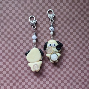 May include: Two cream-colored dog charms with black ears, each attached to a silver-toned lobster clasp. The charms feature white dice beads, small white pearls, and button accents. One dog has a black button eye and a white button on its chest.