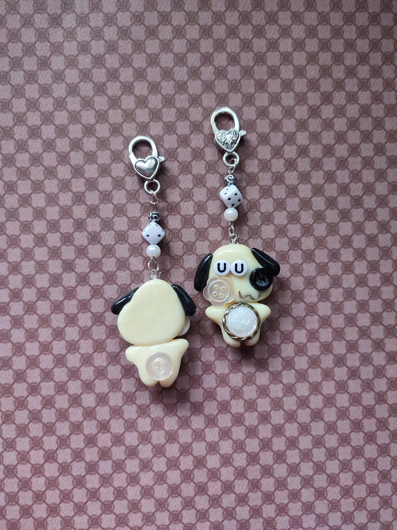 May include: Two cream-colored dog charms with black ears, each attached to a silver-toned lobster clasp. The charms feature white dice beads, small white pearls, and button accents. One dog has a black button eye and a white button on its chest.