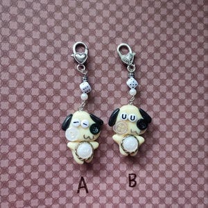 May include: Two cream-colored dog charms with black ears and button eyes, labeled A and B. Each charm features a small white die bead, a silver-tone heart-shaped lobster clasp, and a central white circular accent on the chest area.
