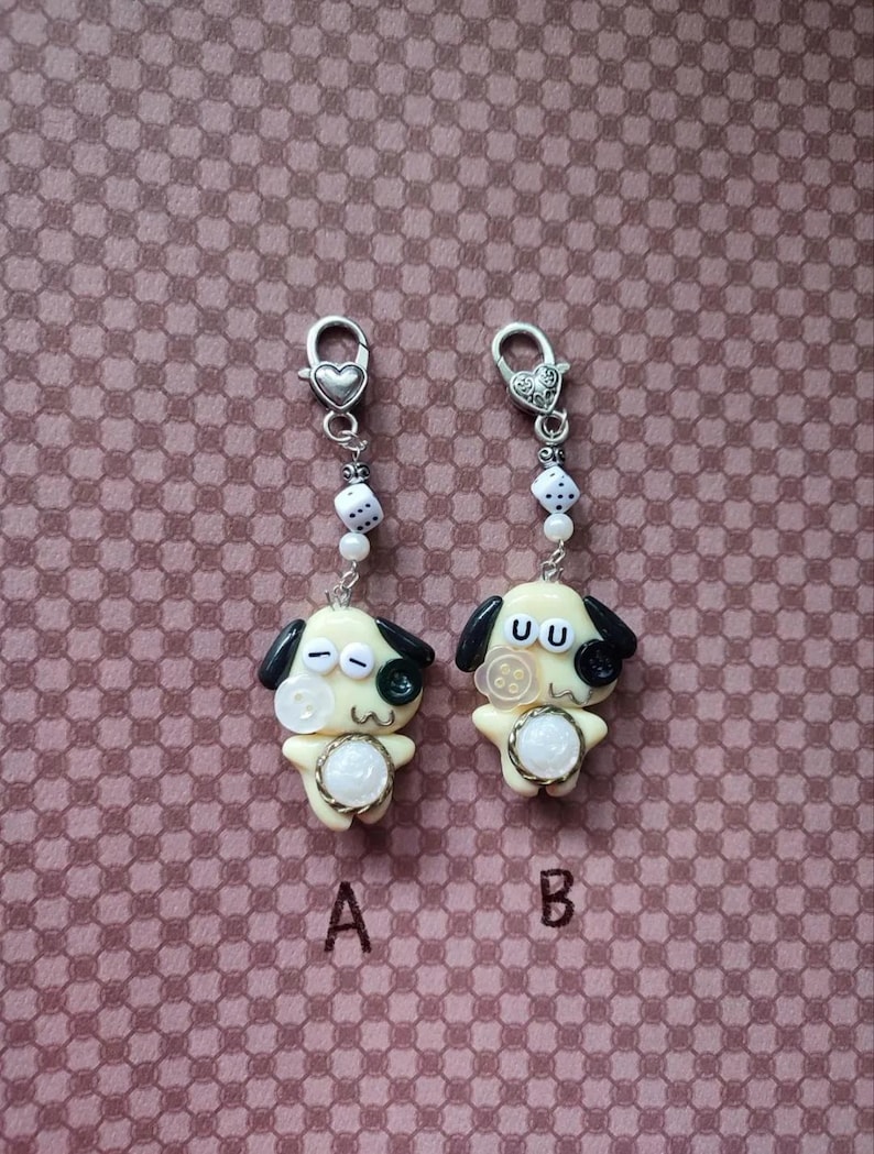 May include: Two cream-colored dog charms with black ears and button eyes, labeled A and B. Each charm features a small white die bead, a silver-tone heart-shaped lobster clasp, and a central white circular accent on the chest area.