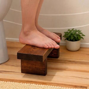 Peut inclure: Un marchepied rectangulaire en bois avec une finition teintée brun foncé et une texture de grain de bois visible. Le marchepied présente une surface supérieure plate soutenue par deux panneaux latéraux verticaux pour les pieds.
