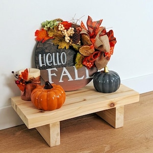 May include: A round wooden sign with the text Hello Fall, decorated with artificial autumn leaves, berries, pinecones, and a plaid ribbon. Beside it sits a small orange ceramic pumpkin, a dark grey pumpkin, and a candle with fall foliage.