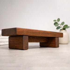 May include: A rectangular wooden riser with a dark brown stained finish and visible wood grain. The platform is supported by two thick, block-shaped legs positioned underneath, creating a low-profile stand with a solid, sturdy construction.