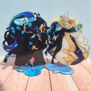 Pode incluir: Dois suportes de acrílico com personagens estilizados em poses dinâmicas. A figura da esquerda tem cabelo azul escuro e um traje preto com motivos de olhos. A figura da direita veste uma túnica branca e dourada com um chapéu alto.