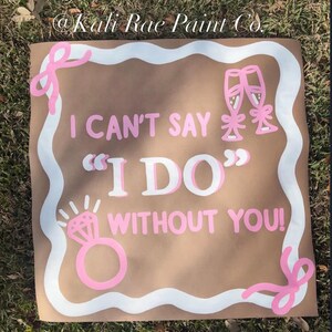 Puede incluir: Un letrero cuadrado marrón con un borde blanco ondulado y detalles de lazos rosas. Presenta el texto "I CAN'T SAY "I DO" WITHOUT YOU!" en blanco y rosa, acompañado de ilustraciones de dos copas de champán y un anillo de compromiso.