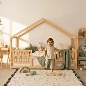 May include: A light-coloured wooden house-shaped bed frame with vertical slats, featuring a mattress, a green star-shaped pillow, a green blanket, and a small stuffed rabbit. A child stands in front of the bed on a star-patterned rug with wooden blocks.