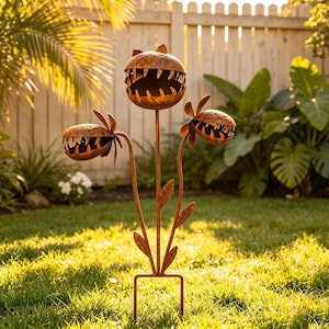 May include: A metal garden stake featuring three stylized plant heads with open, toothy mouths. The sculpture has a weathered, rusted brown finish and is supported by a thin, branched stem structure with several flat, pointed metal leaves attached.