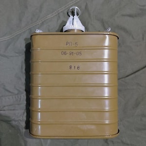 Pode incluir: Um recipiente metálico retangular de cor verde-oliva com nervuras horizontais e um bocal de plástico branco no topo. A superfície frontal apresenta o texto cirílico "РП-5", a sequência numérica "06-91-05" e o número "918" impresso em tinta preta.