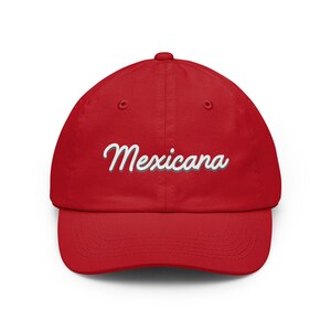 Puede incluir: Una gorra de béisbol de color rojo brillante con la palabra "Mexicana" bordada en una fuente cursiva blanca con un sutil efecto de sombra gris. La gorra tiene una corona estructurada, una visera curva y un botón en la parte superior central.