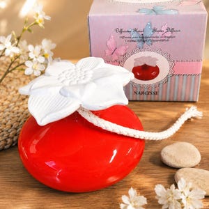 Peut inclure: Un diffuseur d'huiles essentielles en céramique rouge avec un embout en forme de fleur blanche et une mèche en coton blanc. Le diffuseur est accompagné d'une boîte en carton violet clair avec des papillons et le texte Diffuseur Floral Flower Diffuser.