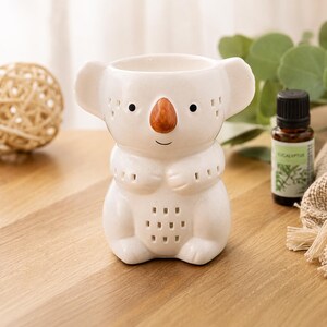 Peut inclure: Un brûleur d'huile en céramique de couleur crème en forme de koala, doté d'un nez marron, d'yeux noirs et de petites découpes carrées sur les oreilles, les bras et le ventre. Le koala présente une ouverture supérieure creuse pour l'huile et la cire.