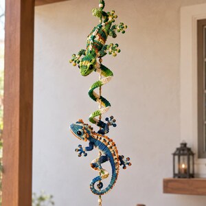 Könnte beinhalten: Ein vertikales Hängemobil mit zwei gehäkelten Geckos, die an einer zentralen Spiralstruktur befestigt sind. Der obere Gecko ist grün und gelb, der untere Gecko ist blau und orange. Ein klarer, facettierter Kristalltropfen hängt ganz unten.