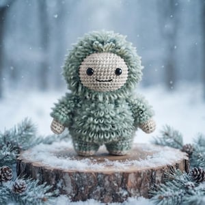 May include: A small, round, sage green crocheted plush toy with a textured, shaggy body and a smooth, light beige face. The figure features two small black bead eyes and a simple embroidered horizontal line for a mouth, standing upright.