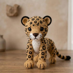 Könnte beinhalten: Eine gehäkelte Leopardenfigur mit beigefarbenem Körper, dunkelbraunen Flecken, weißer Brust und schwarz umrandeten Ohren. Das Stofftier hat große schwarze Augen, eine dunkelbraune Nase und gestickte Schnurrhaare und steht auf vier Beinen.