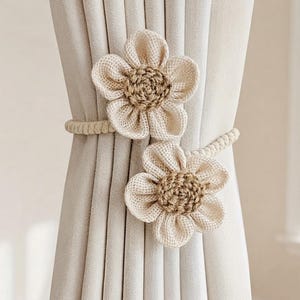 May include: A beige curtain tieback featuring two fabric flowers with textured cream petals and brown crocheted centers. The flowers are attached to a braided cord that wraps around a pleated, light beige curtain panel to hold it in place.