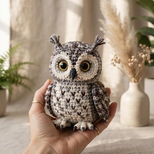 May include: A small, handcrafted crochet owl figurine featuring a textured pattern of grey, white, and brown yarn. The owl has large, round, glossy yellow eyes, a dark grey beak, small tufted ear feathers, and detailed stitching on its chest.