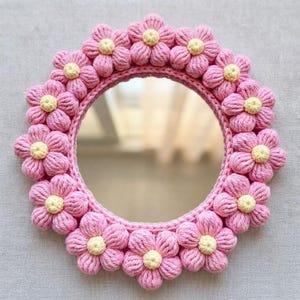 May include: A round mirror framed by a border of crocheted flowers. Each flower features five thick, textured pink petals surrounding a small, pale yellow centre. The flowers are arranged in a continuous ring around the circular glass surface.
