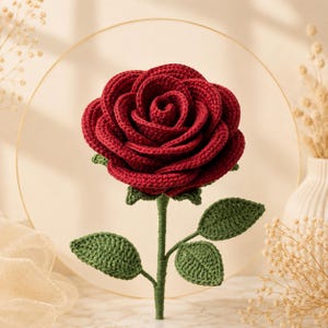 May include: A handcrafted crochet rose featuring deep red, layered petals arranged in a full bloom. The flower is attached to a green stem with three distinct, textured green leaves extending from the sides. The construction shows visible yarn stitches.
