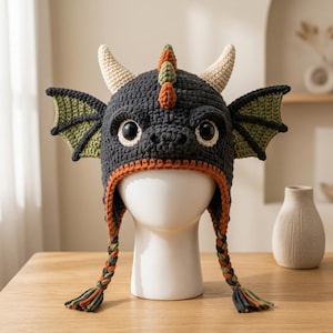 May include: A dark gray crocheted dragon hat featuring two cream-colored horns, green wings on the sides, and a row of orange and green spikes along the top. The hat includes an orange trim and two long, braided tassels in orange, green, and gray.