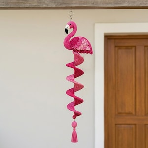 Puede incluir: Una decoración de ganchillo colgante que presenta un flamenco rosa en la parte superior con un pico de punta negra. Debajo del flamenco, una larga cola de ganchillo rosa en espiral se extiende hacia abajo, terminando en una pequeña esfera y borla.