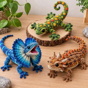 Puede incluir: Tres figuras de lagartos de ganchillo: un gecko verde con manchas naranjas, un lagarto de cuello con volantes azul con la boca abierta y un lagarto cornudo marrón con púas texturizadas. Cada figura tiene grandes ojos oscuros y puntadas detalladas.