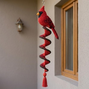 Op de afbeelding: Een gehaakt kardinaalvogel ornament met een felrood lichaam, een zwart gezichtsmasker en een gele snavel. De vogel is bevestigd aan een rood en donkerbruine spiraalvormige staart die verticaal hangt en eindigt in een kleine gehaakte bol en kwast.