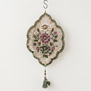 May include: A decorative wall hanging featuring an ornate, quatrefoil-shaped frame in shades of sage green and cream. The interior contains a detailed floral arrangement with white, mauve, and green blossoms, finished with a matching green tassel.