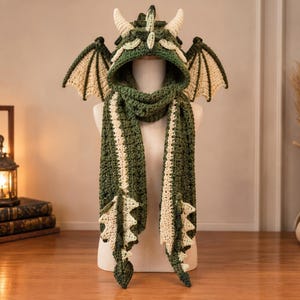 May include: A dark green crochet dragon hood with a long scarf. The hood features two cream-colored horns, a central spine ridge, and two large, cream-colored dragon wings on the sides. The scarf ends have cream-colored triangular spikes.