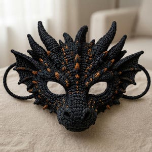 Può includere: Una maschera da drago nera con una superficie testurizzata simile a scaglie e riflessi arancioni sulle creste e sulle corna. La maschera presenta due ali laterali, un muso centrale, due aperture per gli occhi e anelli neri su entrambi i lati.