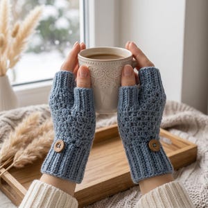 May include: A pair of dusty blue fingerless gloves featuring a textured crochet stitch pattern, a ribbed cuff, and a single round wooden button detail on the outer wrist of each glove, held together around a ceramic mug.