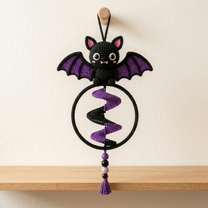 May include: A crocheted black bat ornament with purple wing accents, large white eyes, and pink inner ears. The bat sits atop a black circular hoop containing a purple and black spiral element, finished with a dangling purple tassel and beads.