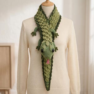 Puede incluir: Una bufanda de ganchillo con forma de lagarto verde con un patrón de escamas texturizado, bordes verde oscuro, grandes ojos negros y una pequeña lengua rosa. El cuerpo del lagarto se extiende por la bufanda, con cuatro patas y una cola larga.