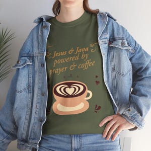 Puede incluir: Una camiseta verde oliva con el texto Jesus & Java powered by prayer & coffee en color beige. Debajo del texto hay un gráfico de una taza de café beige con un diseño de arte latte en forma de corazón. La camiseta se lleva bajo una chaqueta vaquera azul claro.