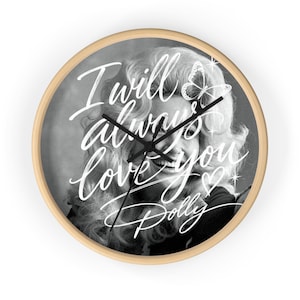 Op de afbeelding: Een ronde wandklok met een lichthouten frame met een zwart-wit portret van Dolly Parton. De tekst I will always love you Dolly staat in witte letters over de afbeelding geschreven, vergezeld door een vlinder en een klein hartje.