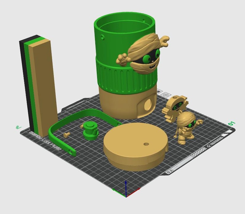 以下が含まれることがあります： 3Dプリントされた緑とベージュの円筒形容器。緑色の目をしたミイラのキャラクター、同色のベージュの蓋、緑色のハンドル、2体の小さなミイラのフィギュアが含まれています。各パーツは黒いグリッド状のプリントベッド上に配置されています。