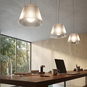 May include: Three pendant lights with clear, ribbed, conical shades hang from the ceiling. Each fixture features a visible internal light bulb, casting a warm glow over a wooden desk equipped with a laptop, a ceramic mug, and various office supplies.