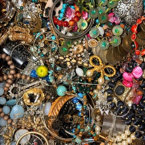 May include: A large collection of assorted jewelry including gold-tone chains, beaded necklaces, metal bangles, rhinestone-encrusted pieces, colorful plastic charms, faux pearls, and various metallic pendants in a dense, overlapping arrangement.